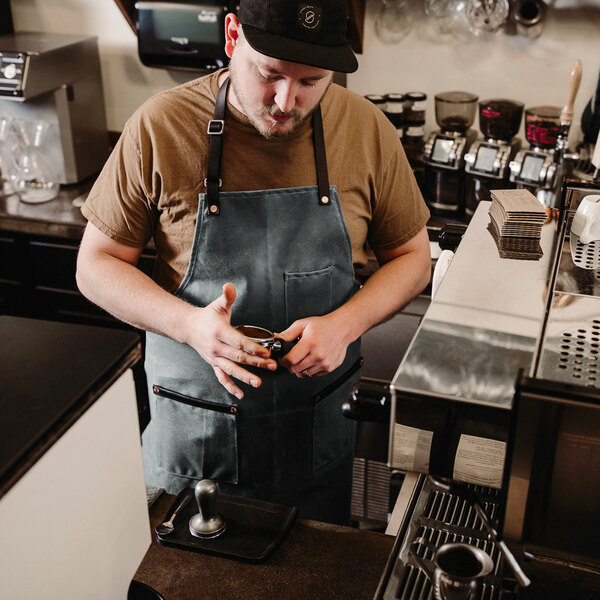 A man in a black Hardmill waxed canvas bib apron holding a cup of coffee.