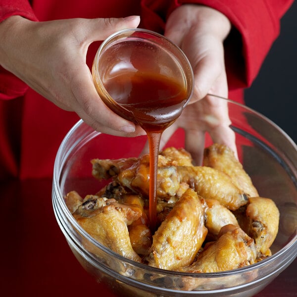A person pouring honey garlic sauce from a small bowl onto cooked chicken wings in a glass mixing bowl.