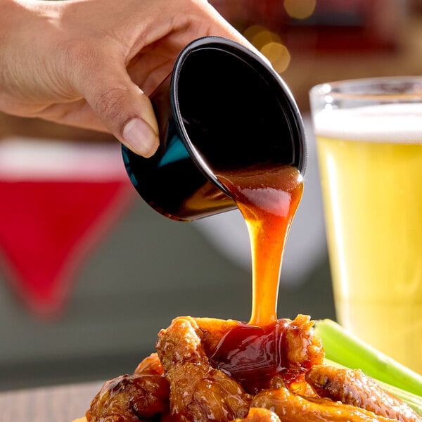 A hand pouring Frank's RedHot Stingin' Honey Garlic Sauce from a small black cup onto a plate of chicken wings, with a glass of beer and celery in the background.