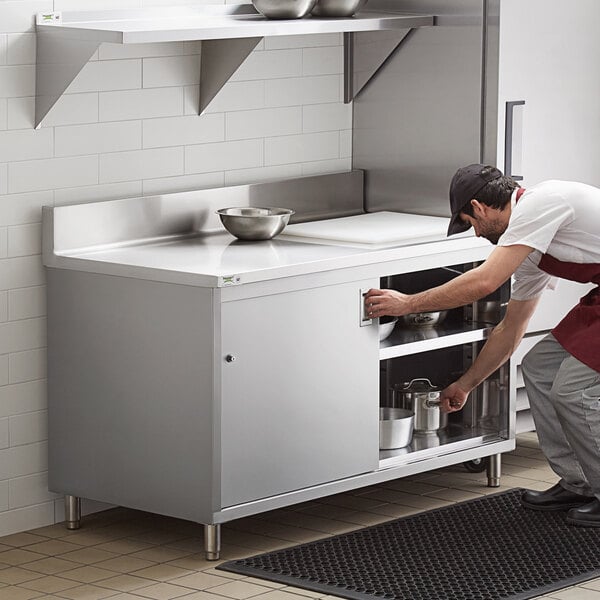 A man in a white shirt and black pants bending over a Regency stainless steel enclosed base table with sliding doors.