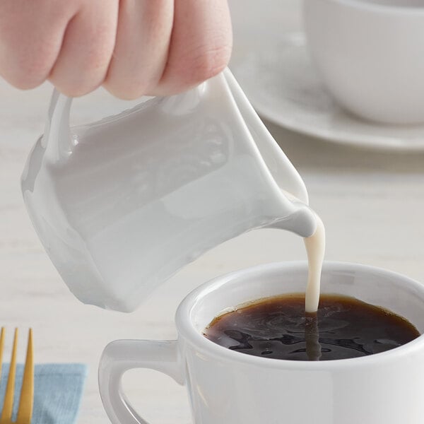 A person pouring milk from a white pitcher into a cup of coffee.