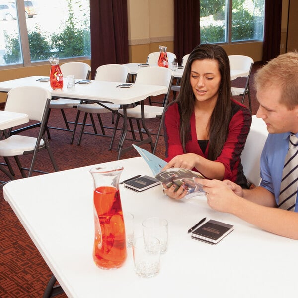 A man and woman sitting at a white Lifetime plastic folding table with a glass on it.