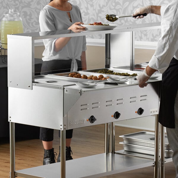 A man and woman serving food from a ServIt electric steam table at an outdoor catering setup.