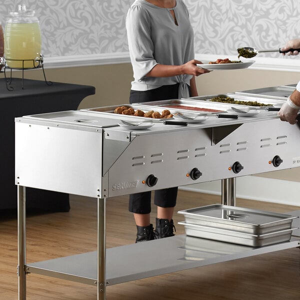 A woman serving food from a ServIt open well steam table to a man and woman.
