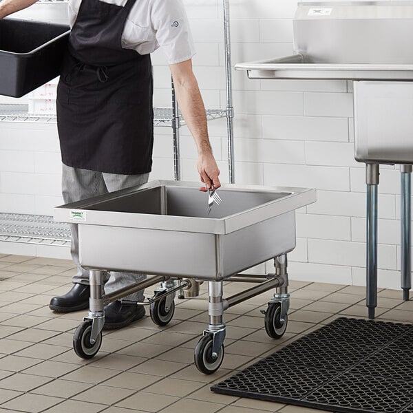 A man in a black apron using a Regency mobile silverware soaking sink on a counter.