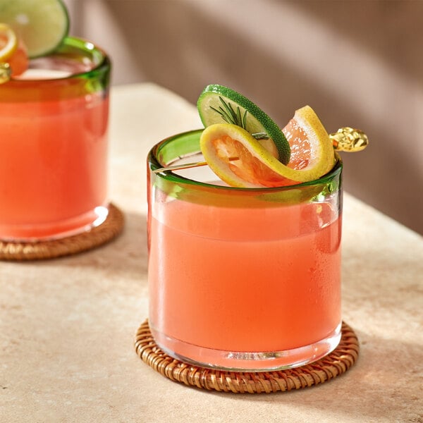Two Acopa Tropic rocks glasses with orange drinks and lime slices on a coaster.