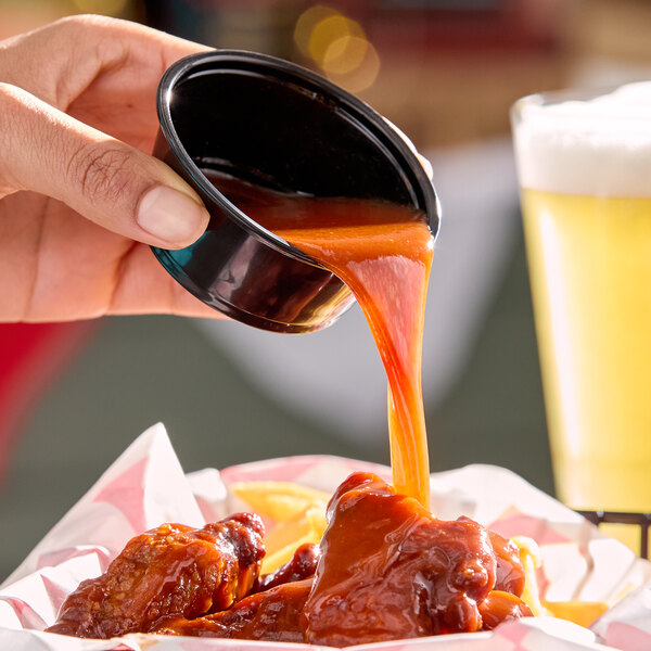 A hand pours bright red hot sauce from a small black cup onto chicken wings in a basket lined with paper.