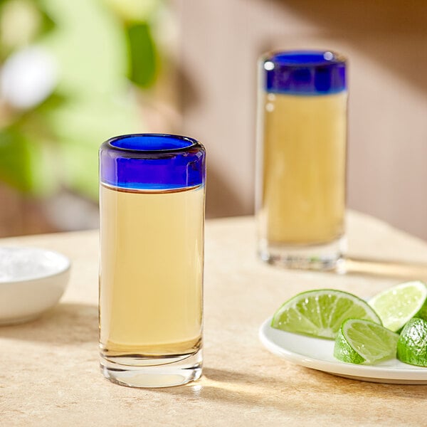 Two Acopa Tropic shooter glasses with a blue rim filled with tequila next to a lime wedge.