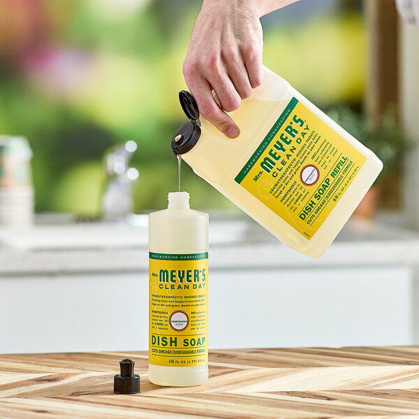 A hand pouring Mrs. Meyer's Honeysuckle Scented dish soap into a plastic bottle.