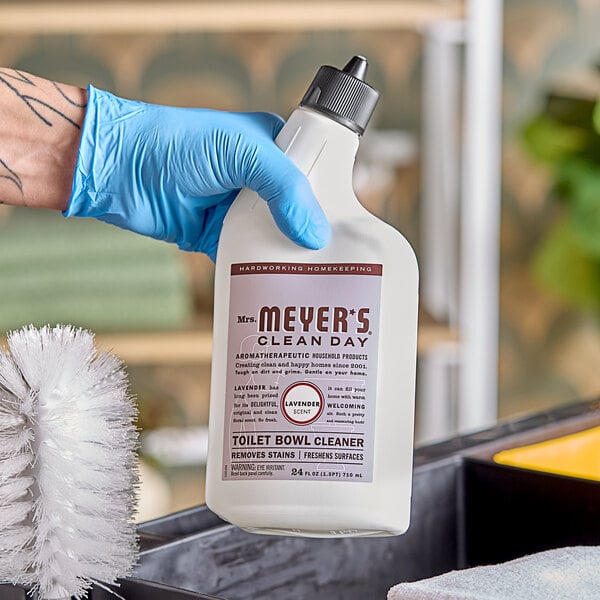 A gloved hand holding a white bottle of Mrs. Meyer's lavender toilet bowl cleaner.