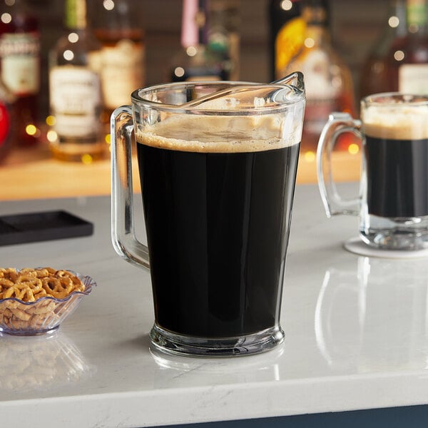 An Arcoroc glass beer pitcher of dark beer on a counter with a bowl of pretzels.