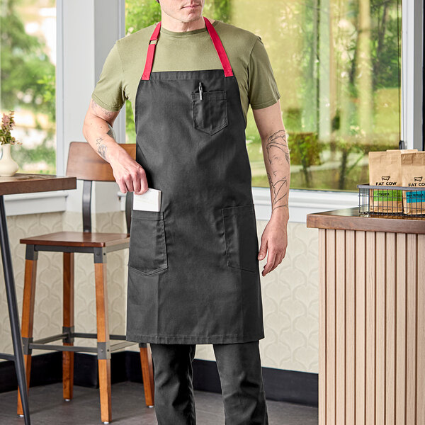 A man wearing a black Uncommon Chef Rebel bib apron with red webbing standing in a professional kitchen.