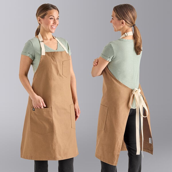 Two women wearing Uncommon Chef caramel bib aprons with natural webbing and pockets.