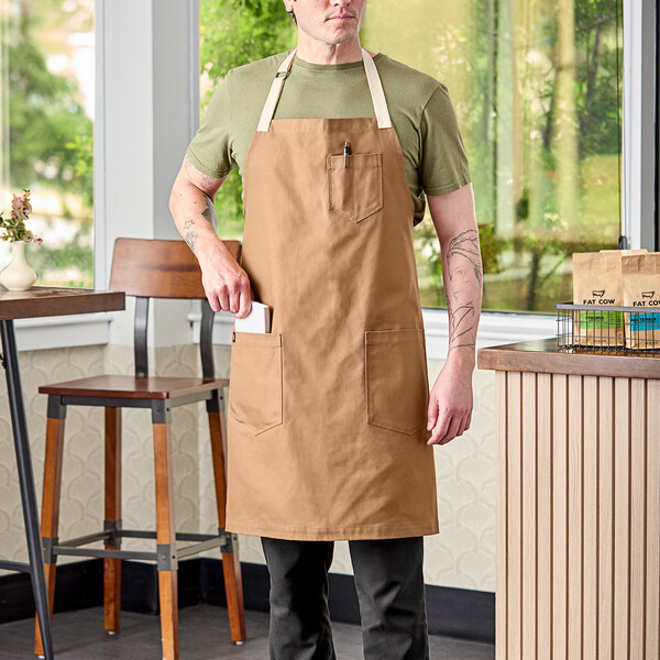 A man wearing a brown Uncommon Chef Rebel apron standing in a professional kitchen.