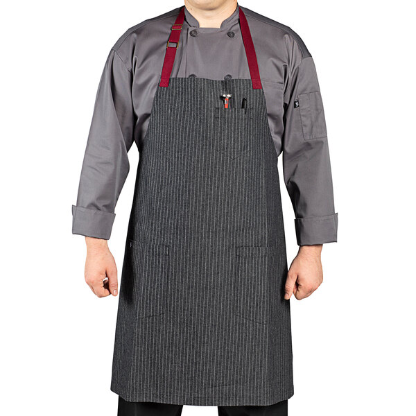 A man wearing a Uncommon Chef pinstripe denim bib apron with burgundy accents and 3 red pockets.