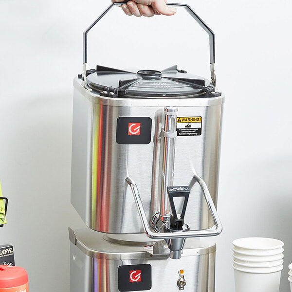 A hand holding a Grindmaster stainless steel coffee shuttle over a stack of white cups.