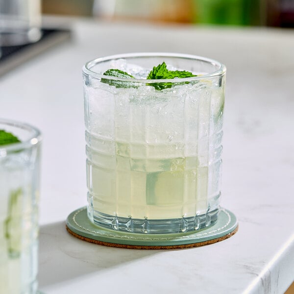 A clear, textured rocks glass filled with a light-colored beverage, ice, and garnished with mint, sitting on a coaster.