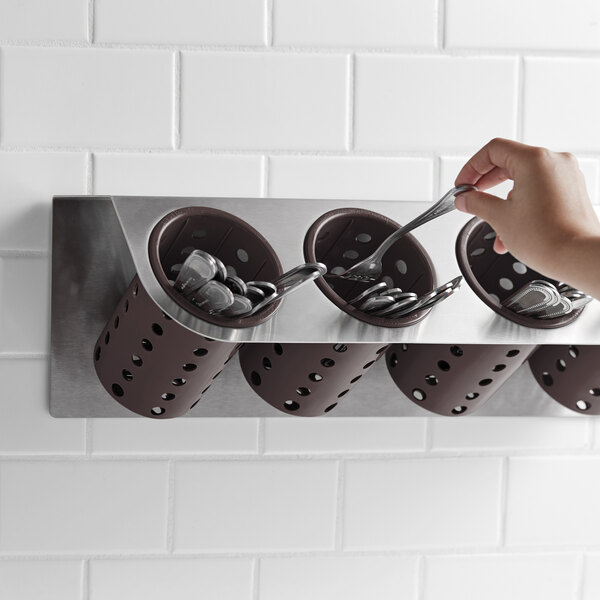 A hand holding a spoon in a Steril-Sil stainless steel flatware organizer with brown plastic cylinders.