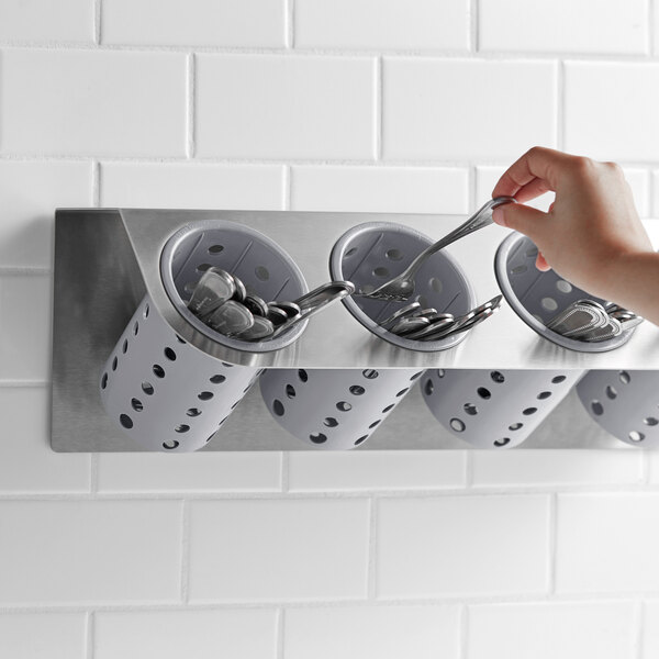 A hand holding a spoon in a Steril-Sil stainless steel flatware holder with gray plastic cylinders.