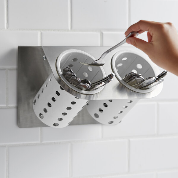 A hand holding a spoon in a Steril-Sil stainless steel flatware holder with white plastic cylinders.