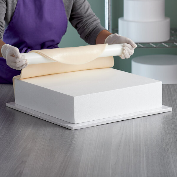 A woman cutting a Baker's Mark foam square cake dummy with a knife.