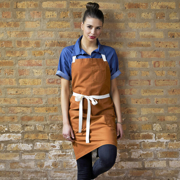 A woman wearing a Uncommon Chef Walnut Canvas Bib Apron with natural webbing and 3 pockets leaning against a brick wall.