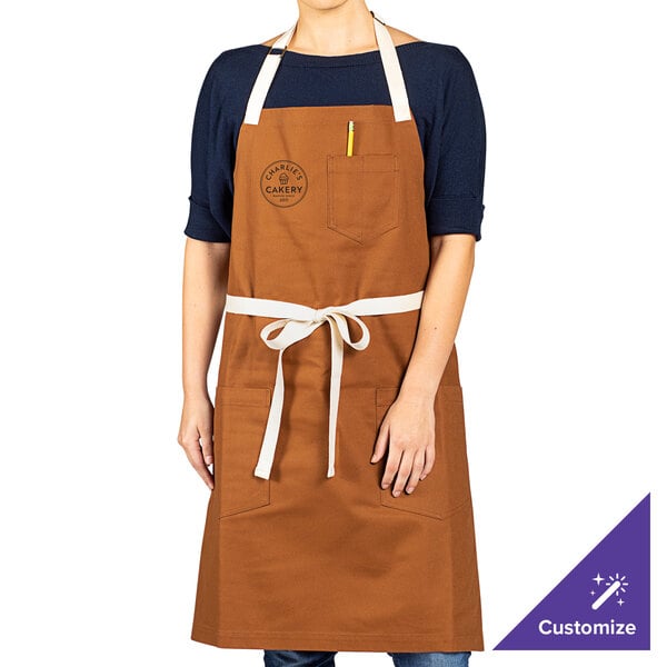 A brown cotton canvas bib apron with natural webbing straps and three pockets, shown being worn by a person.