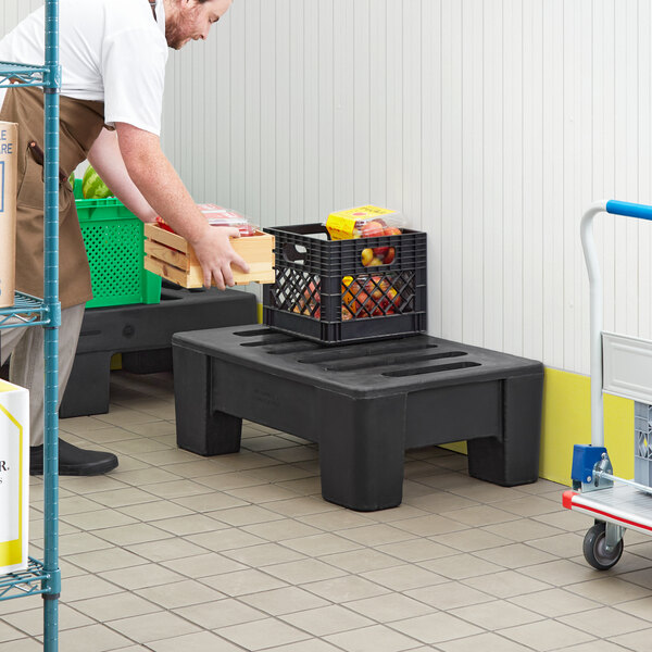 A man in a white shirt and brown pants using a black plastic Regency Dunnage Rack to carry crates.