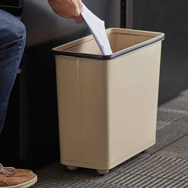 A person's hand putting paper into a beige Rubbermaid wastebasket.