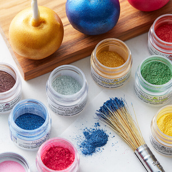 A jar of Roxy & Rich Almond Sparkle Dust in gold on a table with other jars of colorful powder.