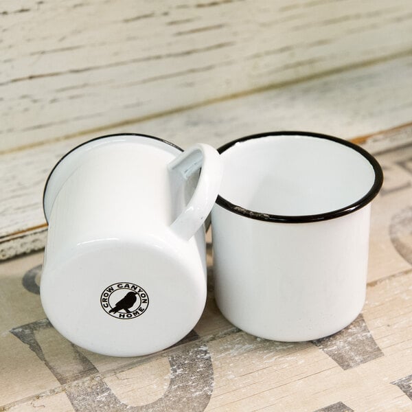 Two white Crow Canyon Home enamelware mugs with black rims on a table.