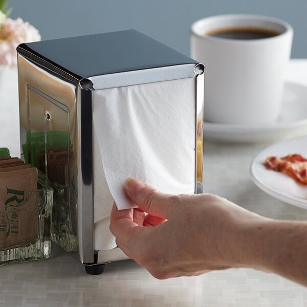 A hand pulling a Choice White Junior low-fold dispenser napkin out of a dispenser on a counter in a breakfast diner.