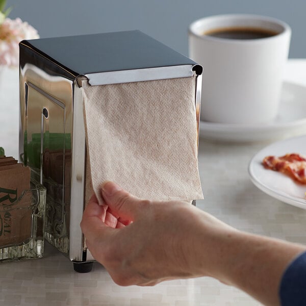 A metal napkin dispenser with a kraft natural low-fold napkin being pulled out by a hand.