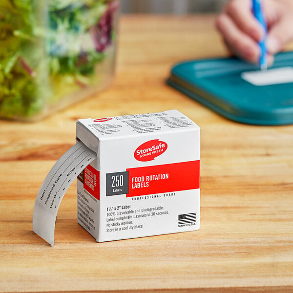 A small white box of Cambro StoreSafe labels on a wooden surface with a hand holding a blue pen