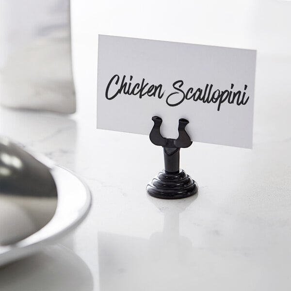 An American Metalcraft black metal low profile harp card holder on a counter with a place card in it.