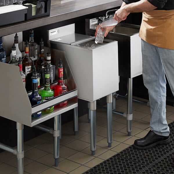 A person is scooping ice from a stainless steel underbar ice bin with a sliding lid, next to a rack of assorted liquor bottles.