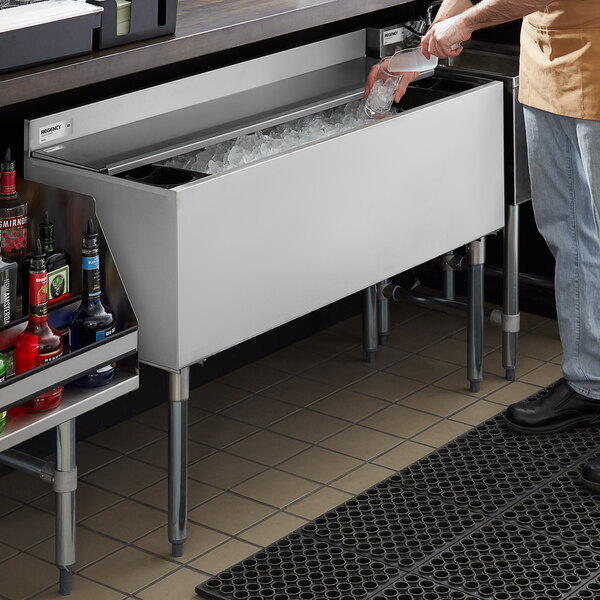 A person is scooping ice from a large stainless steel ice bin located under a bar counter.