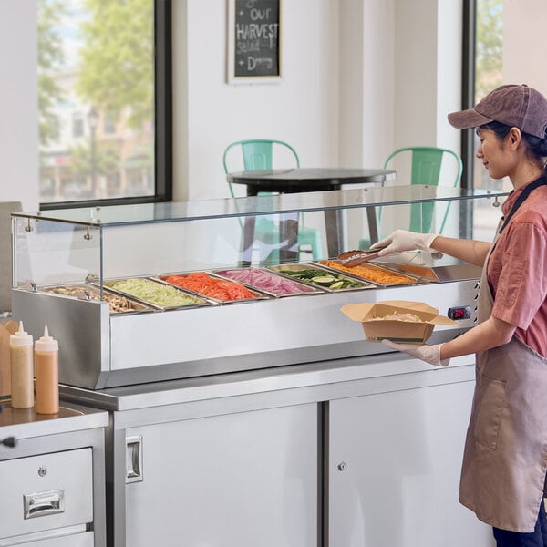 A stainless steel countertop refrigerated prep rail with a sneeze guard, filled with various fresh ingredients and being used by a food service worker.
