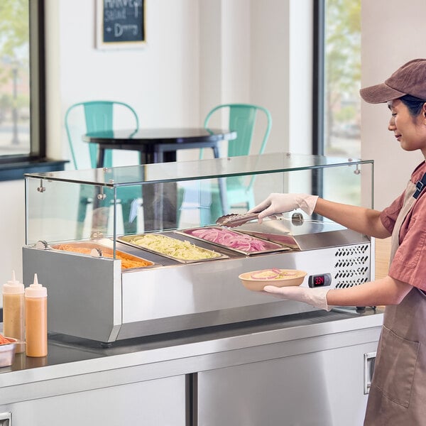 A stainless steel countertop refrigerated prep rail with a glass sneeze guard, containing several food pans and being used by a person serving food.
