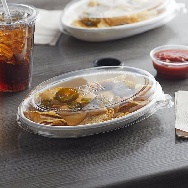 A plastic container of nachos with a red sauce and Eco-Products WorldView oval lid on a table with a drink.