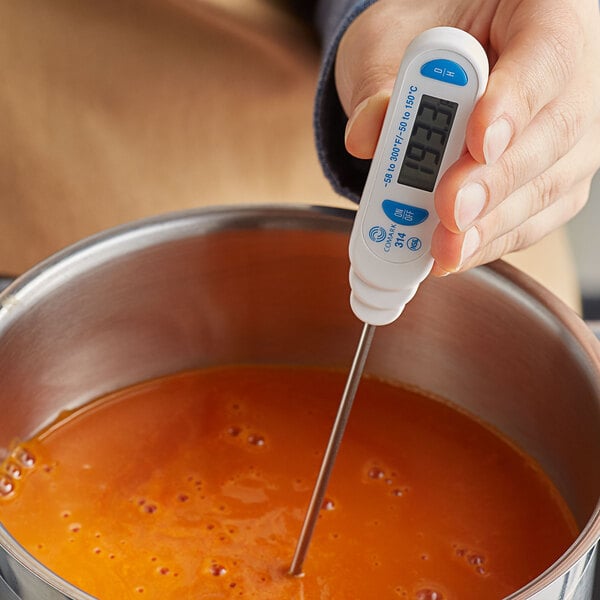 A hand holding a Comark digital pocket probe thermometer over a pot of soup.