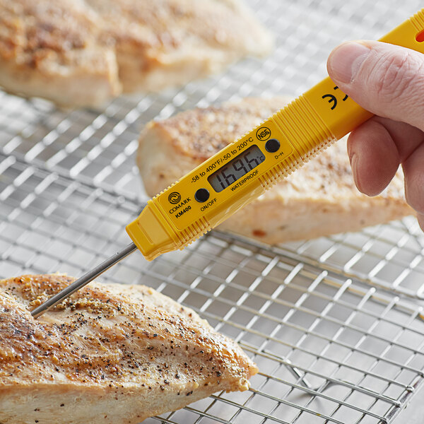 A hand using a yellow Comark digital pocket probe thermometer to check the temperature of meat on a grill.