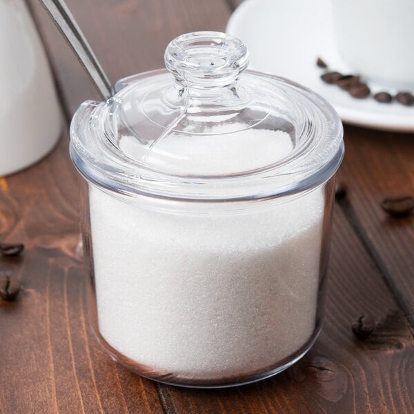 A glass Cambro condiment jar with a slotted lid full of sugar with a spoon.