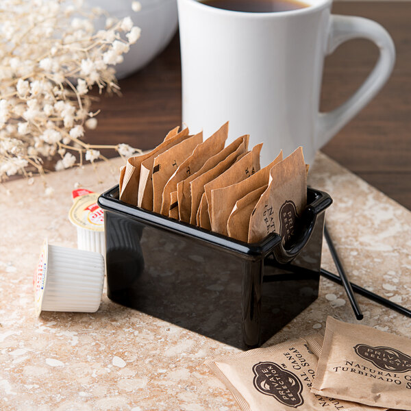 A Carlisle black plastic sugar caddy with brown packets next to a white mug.