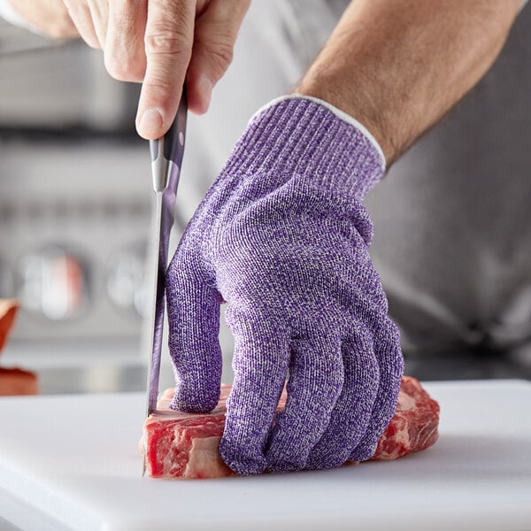 A person cutting meat with a knife while wearing purple San Jamar cut resistant gloves.