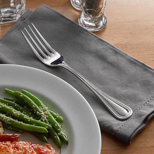 An Acopa stainless steel dinner fork on a napkin next to a plate of food.