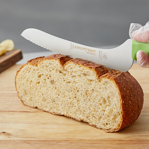 A gloved hand uses a Dexter-Russell green scalloped bread knife to cut a loaf of bread on a cutting board.