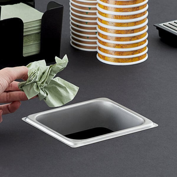 A hand dropping a green napkin into a rectangular stainless steel in-counter trash chute.