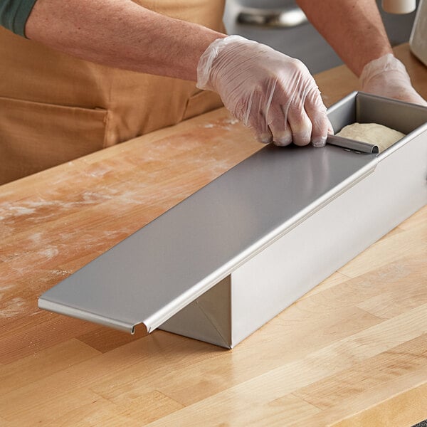 A person wearing gloves slides a metal cover onto a Baker's Mark Pullman bread pan.