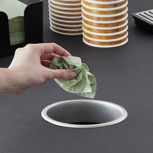 A hand dropping crumpled green paper into a stainless steel in-counter trash chute.